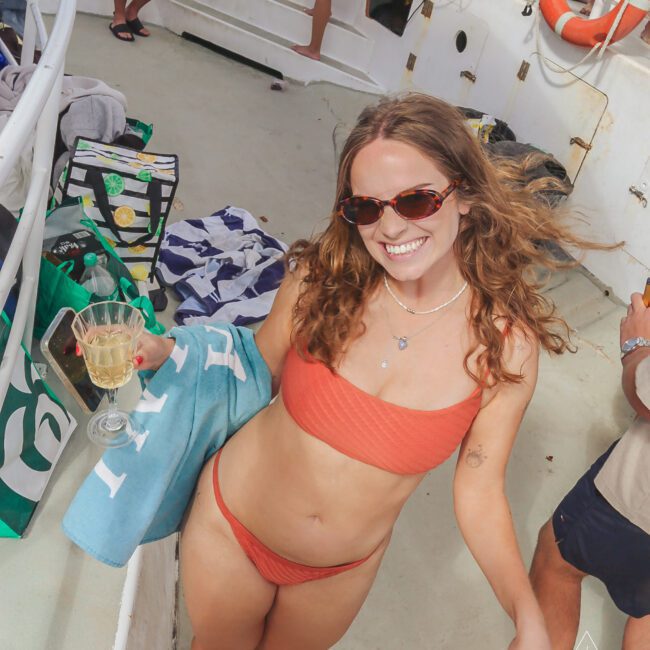 Woman in an orange bikini and sunglasses smiles at the camera, holding a towel and a glass of wine on a boat. Other people relax in the background, with bags and towels visible on the deck.