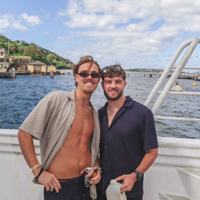 Two men stand on a boat deck by the water, smiling at the camera. One is shirtless under an open shirt, holding a drink, while the other wears a dark shirt and holds a bottle of orange juice. Sunny day with blue sky and clouds.