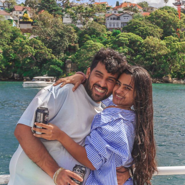 A smiling couple stands by a waterfront, hugging and holding cans, with green trees and houses on a hill in the background under a partly cloudy sky.