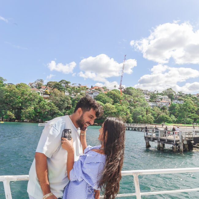 A young couple smiles and laughs together on a boat, with water, a dock, and green trees in the background under a bright blue sky with scattered clouds.