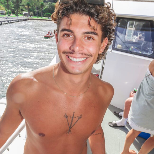 A young man with curly hair and a mustache, wearing sunglasses on his head and a necklace, stands shirtless on a boat, smiling at the camera. There is water and greenery in the background.