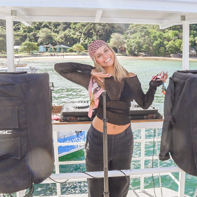 A smiling woman in a black outfit and headscarf poses on a boat between two speakers, with water and a tree-lined shore in the background. She holds sunglasses and keys, with one hand resting on her chin.