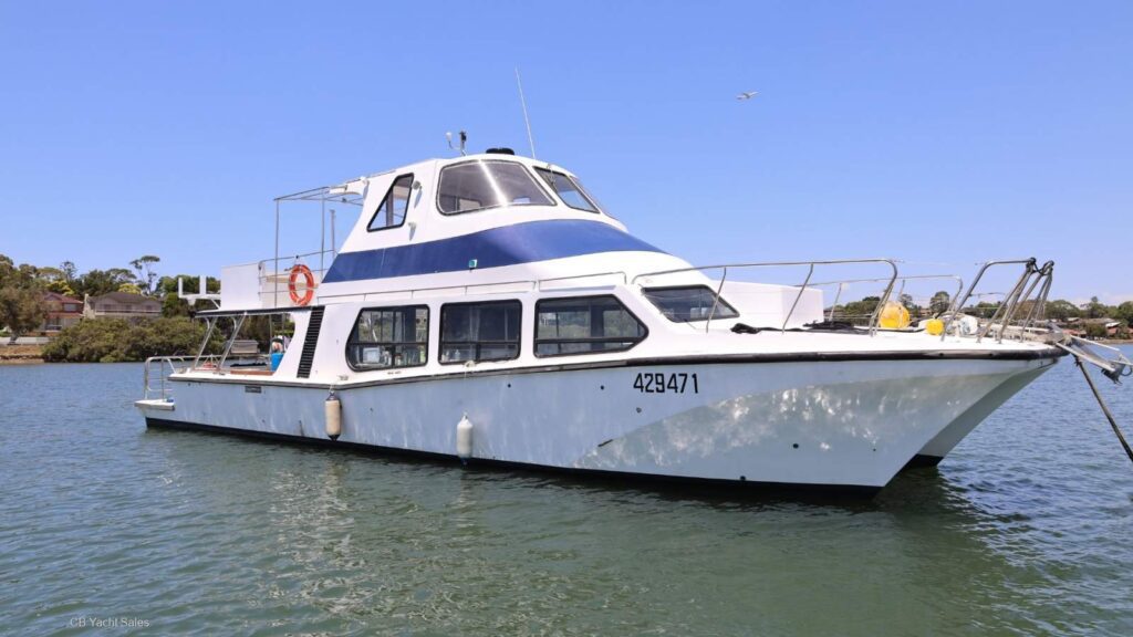 A white and blue passenger boat with large windows floats on calm water near the shore under a clear blue sky. The boat has the number 429471 and features a lifebuoy and outdoor deck area.