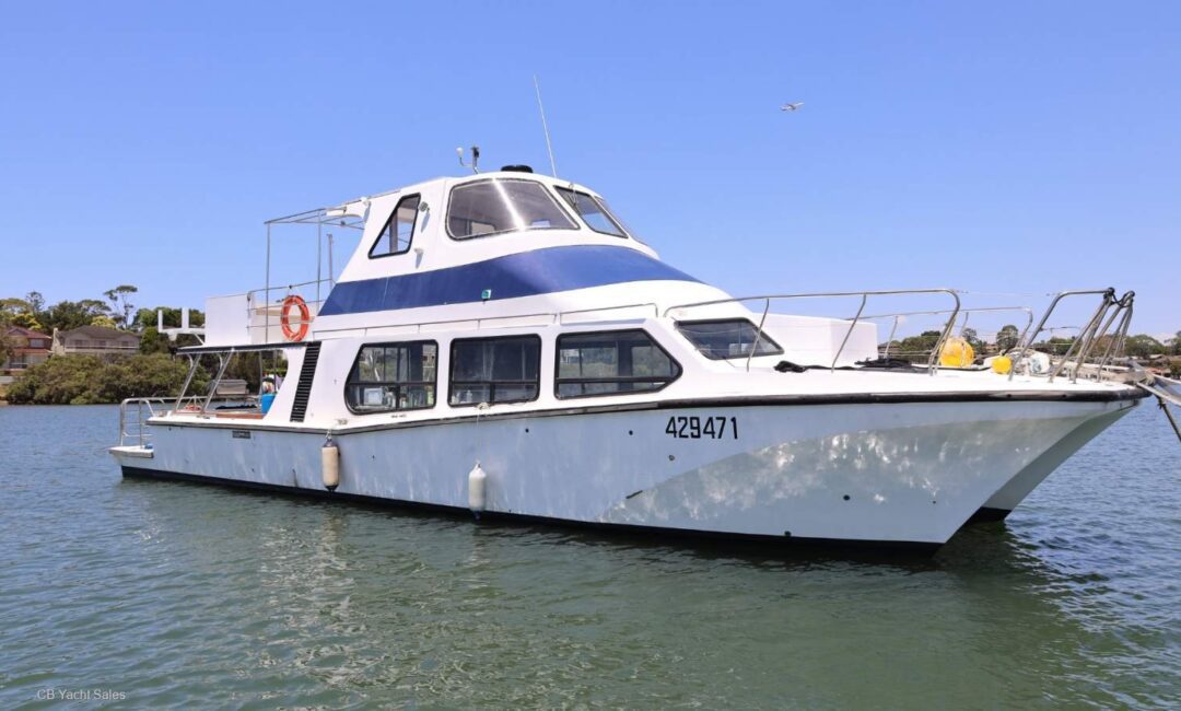 A white and blue passenger boat with large windows floats on calm water near the shore under a clear blue sky. The boat has the number 429471 and features a lifebuoy and outdoor deck area.