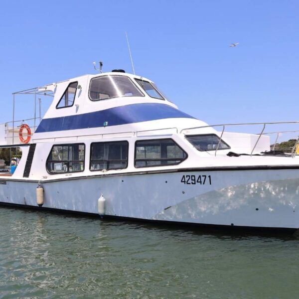 A white and blue passenger boat with large windows floats on calm water near the shore under a clear blue sky. The boat has the number 429471 and features a lifebuoy and outdoor deck area.