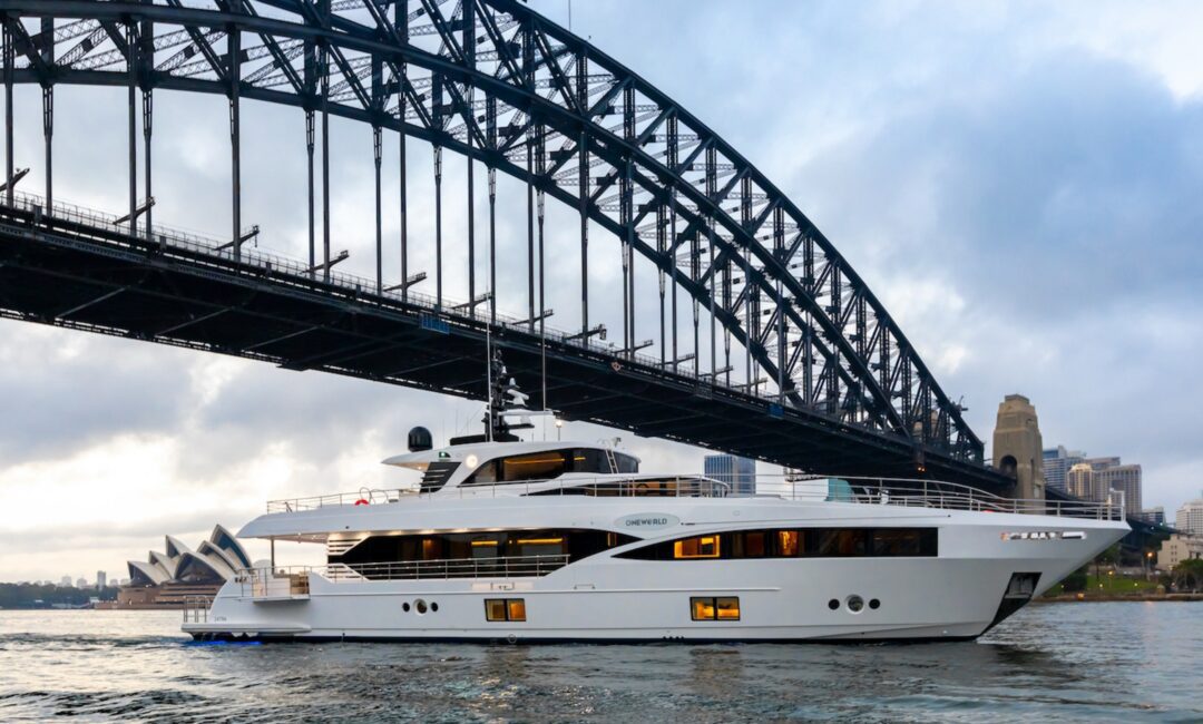 A large white yacht cruises under the Sydney Harbour Bridge with the Sydney Opera House and city skyline visible in the background on a cloudy day.