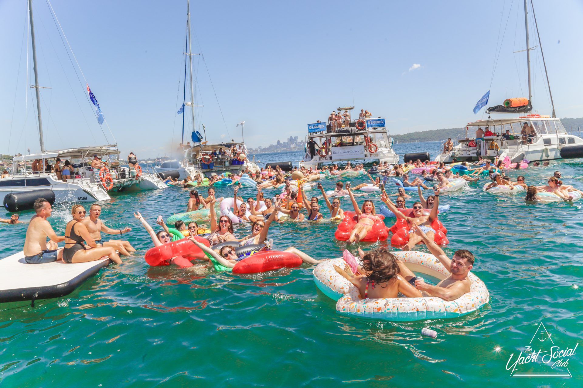 A group of people on colorful inflatables and pool floats enjoy a sunny day in the water near anchored boats during a vibrant Yacht Social, surrounded by blue-green water and a distant city skyline under a clear sky.