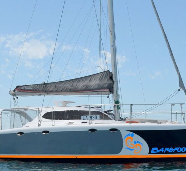 A modern catamaran sailboat with a gray hull and white deck, featuring the name "Barefoot" and an orange foot logo, floating on calm blue water under a partly cloudy sky.