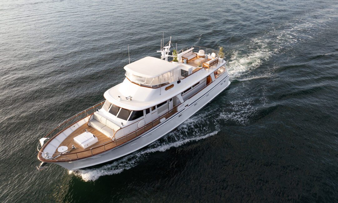 A white motor yacht cruises on calm, open water, creating a gentle wake behind it. The yacht has wooden accents, a shaded upper deck, and appears to be traveling smoothly on a clear day.