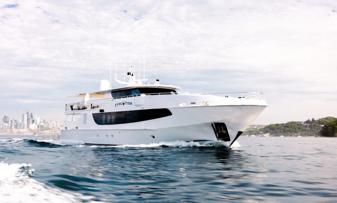 A large white yacht cruises on calm blue water near a city skyline, with green trees visible on the right and a partly cloudy sky overhead.