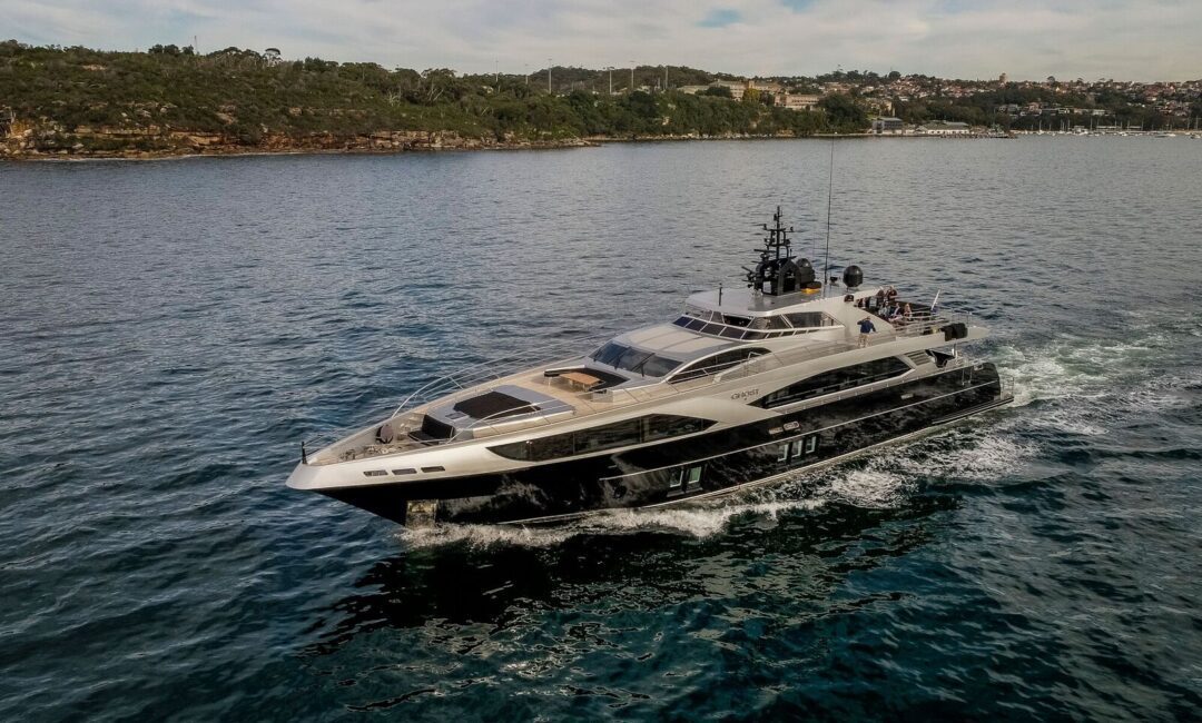 A sleek, modern yacht cruises through calm blue water near a forested shoreline with scattered buildings visible in the distance under a partly cloudy sky.