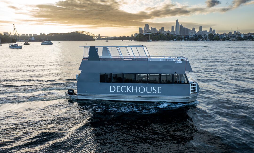 A modern gray boat named DECKHOUSE cruises on a river at sunset, with city buildings and the Sydney Harbour Bridge visible in the background.