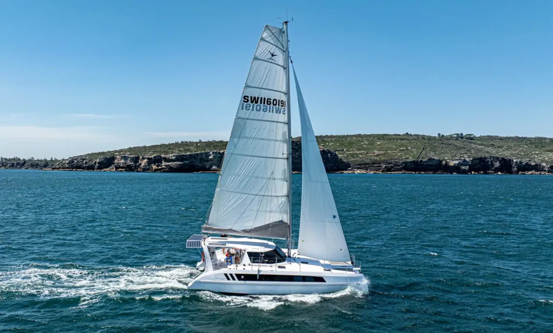 A white sailboat with large sails glides across a blue sea near a rocky coastline under a clear blue sky.
