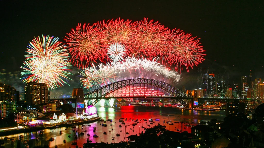 Colorful fireworks light up the night sky above Sydney Harbour Bridge, reflecting on the water during a lively harbour celebration. City buildings and boats are visible, with many enjoying the festivities aboard New Year’s Eve yacht hire, marking a memorable Sydney event.