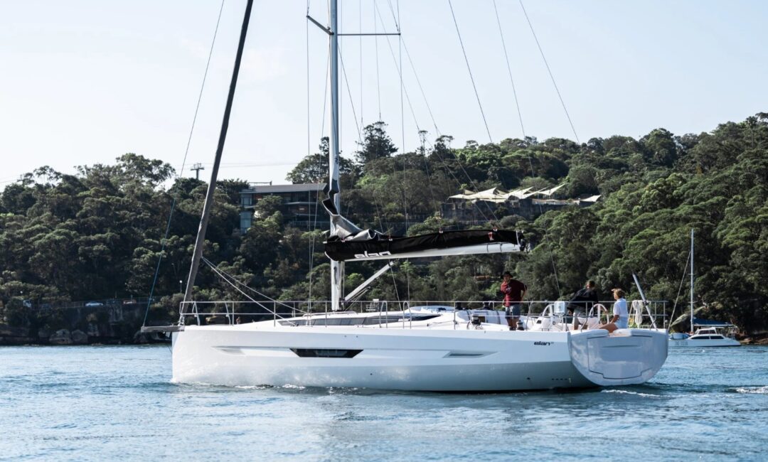 A white sailboat with people onboard sails on calm water near a forested shoreline with buildings visible among the trees under a clear sky.