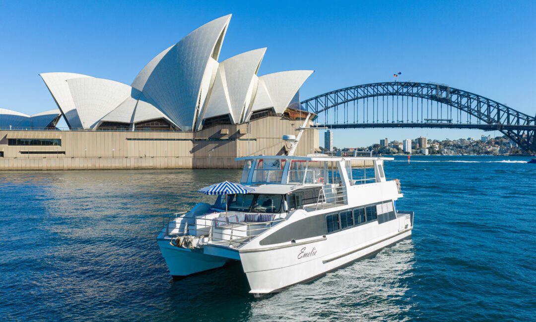 A white boat cruises near the Sydney Opera House and Sydney Harbour Bridge on a sunny day with clear blue skies and calm water.