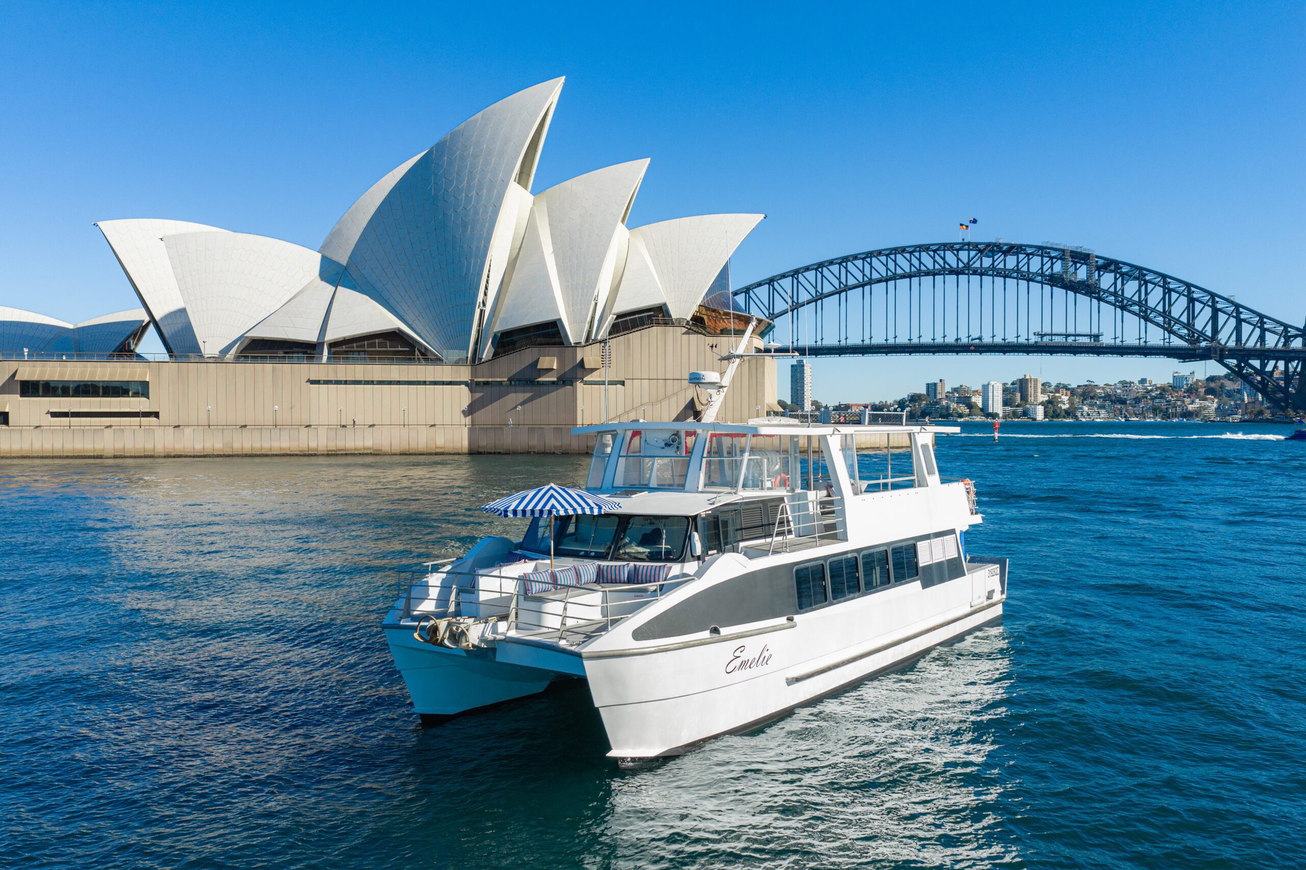 A white boat cruises near the Sydney Opera House and Sydney Harbour Bridge on a sunny day with clear blue skies and calm water.