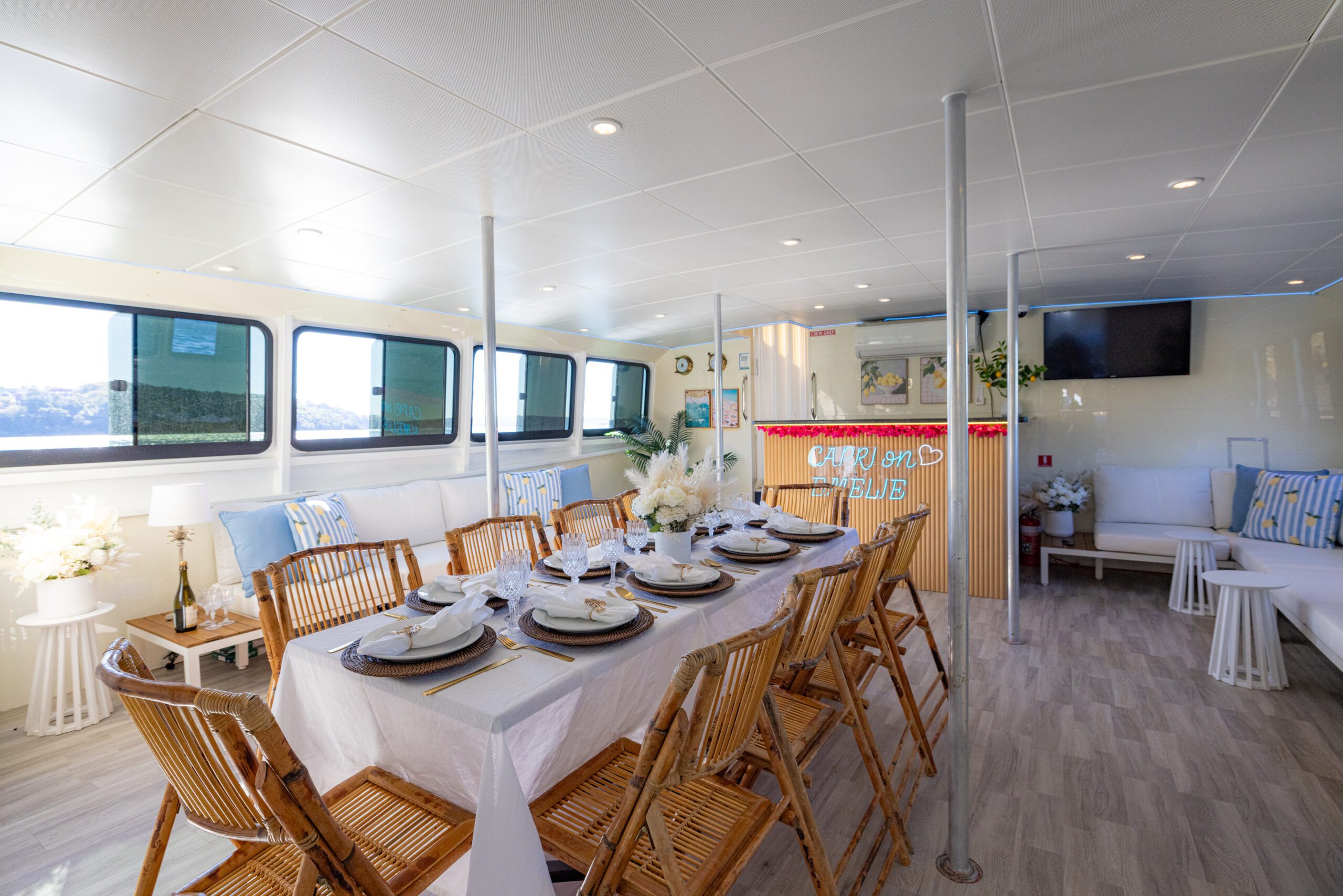 A spacious dining area on a boat, featuring a table set for eight with white linens, wooden chairs, large windows, a bar with neon lights, and cozy lounge seating in a bright, modern interior.