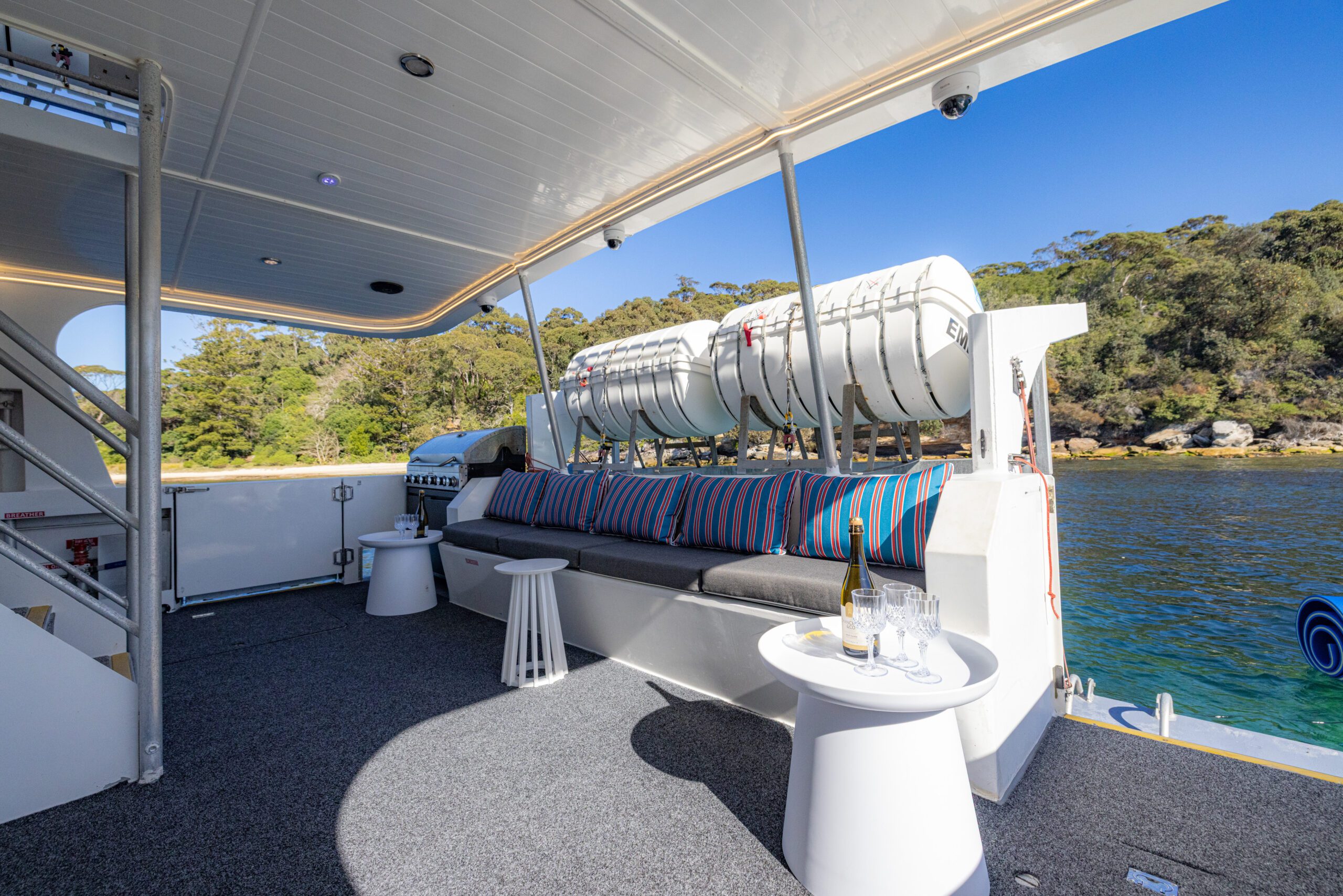 Outdoor yacht deck with cushioned seating, striped pillows, tables with drinks, and life rafts above. Water and trees are visible in the background on a sunny day.