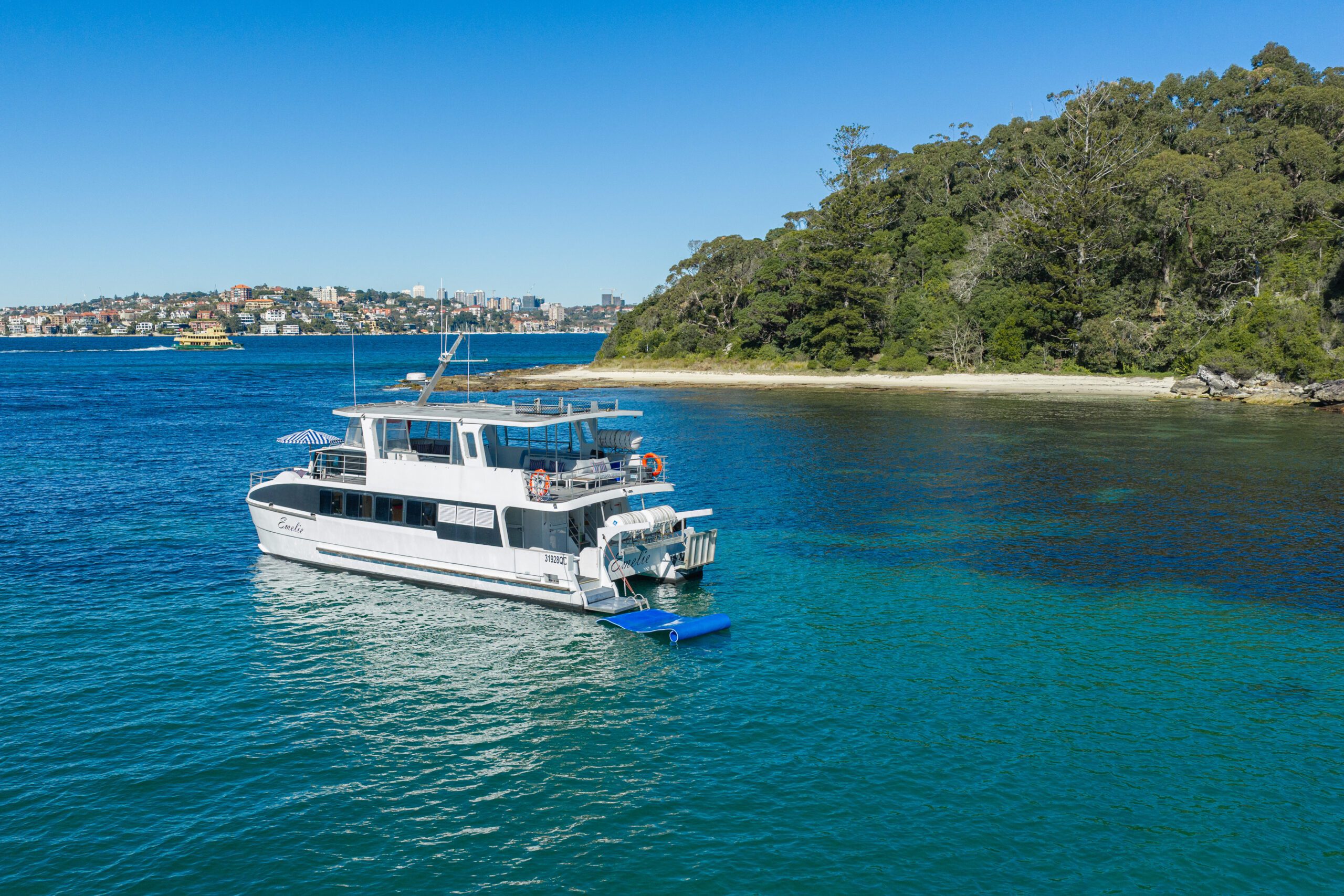 A white yacht floats on clear blue-green water near a tree-lined shoreline, with city buildings visible in the distant background under a clear blue sky.