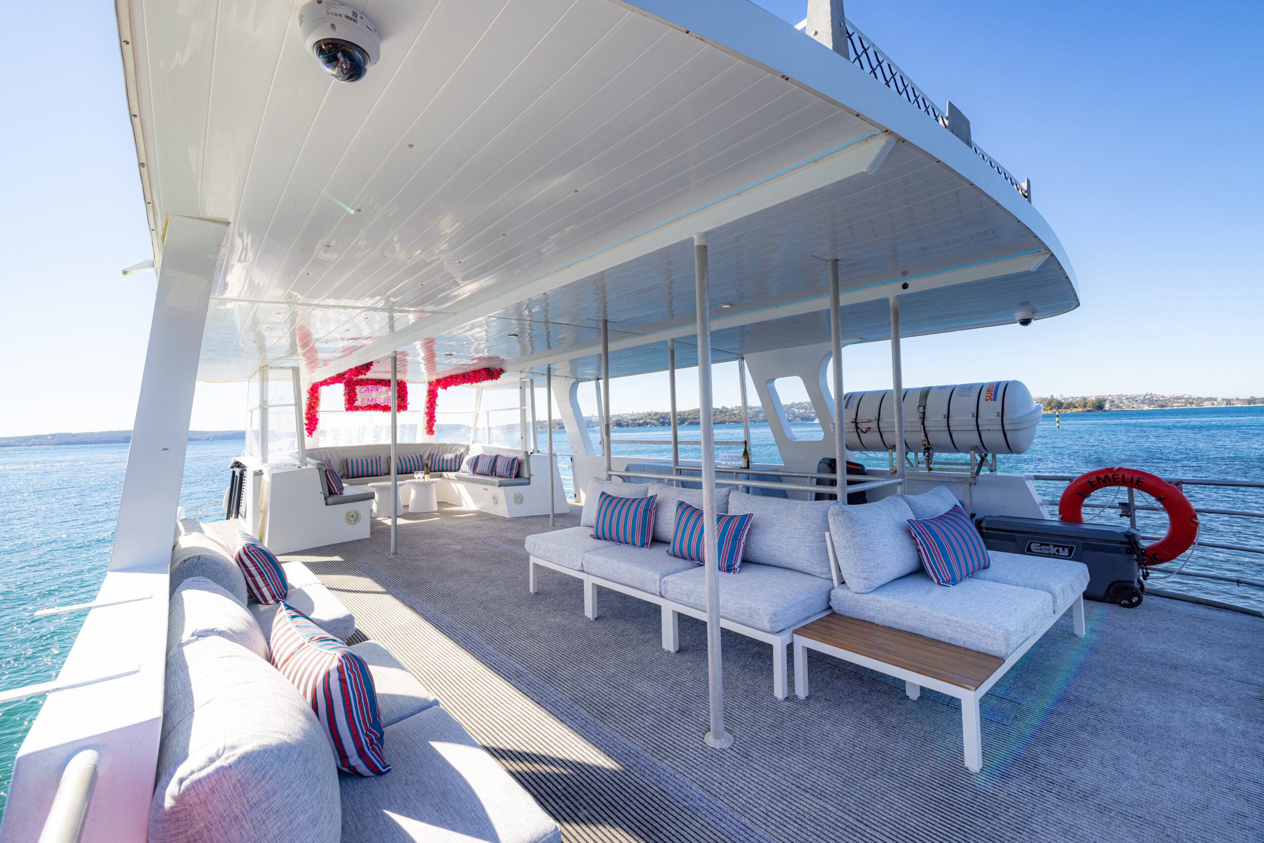 Spacious yacht deck with cushioned lounge seating, striped pillows, and tables, overlooking calm blue water under a clear sky. Red life preserver attached to railing on right.