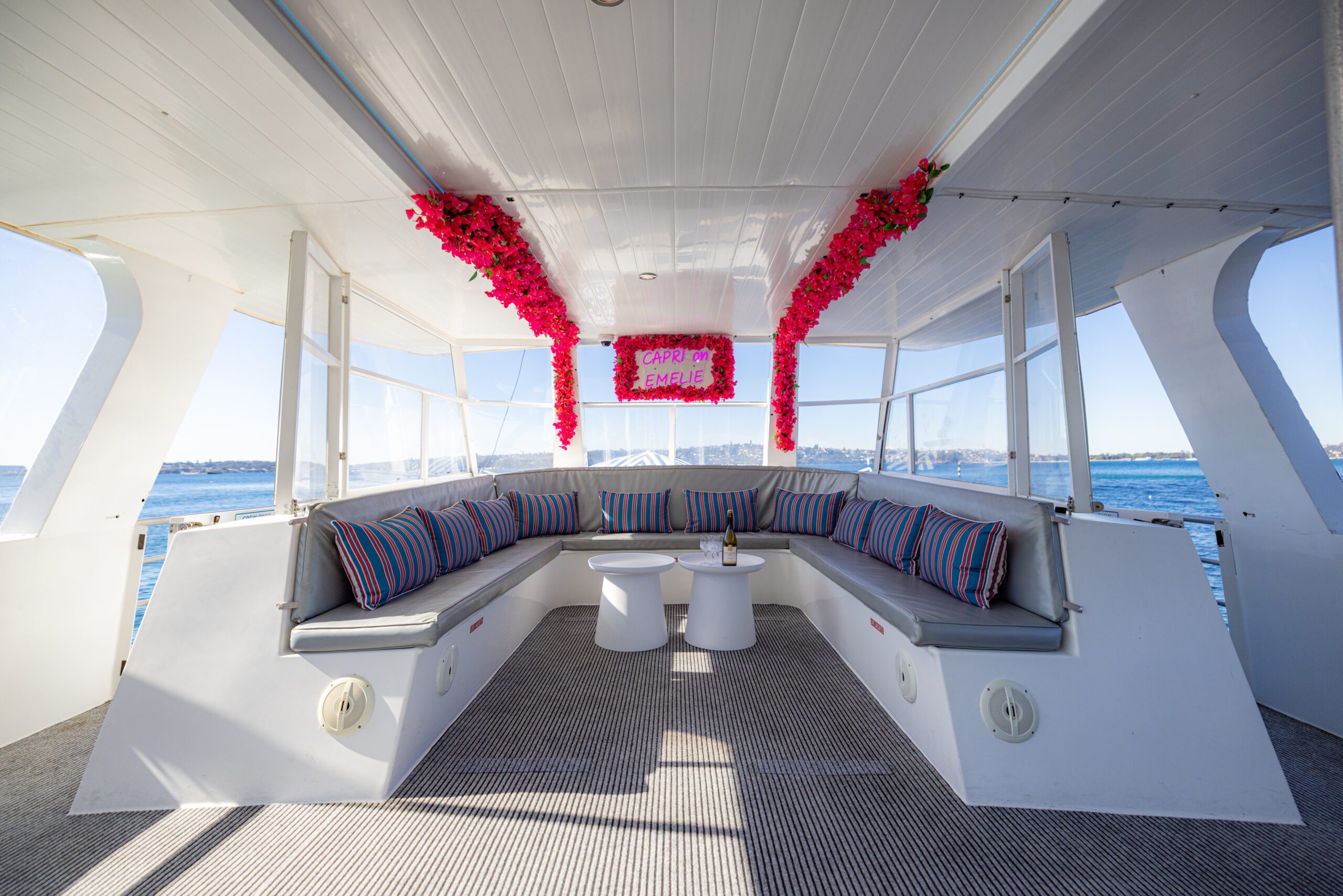 Inside view of a boat with gray bench seating, striped pillows, two small tables, champagne, and pink flowers hanging above a “Happy Birthday” sign. Large windows show blue water and sky outside.