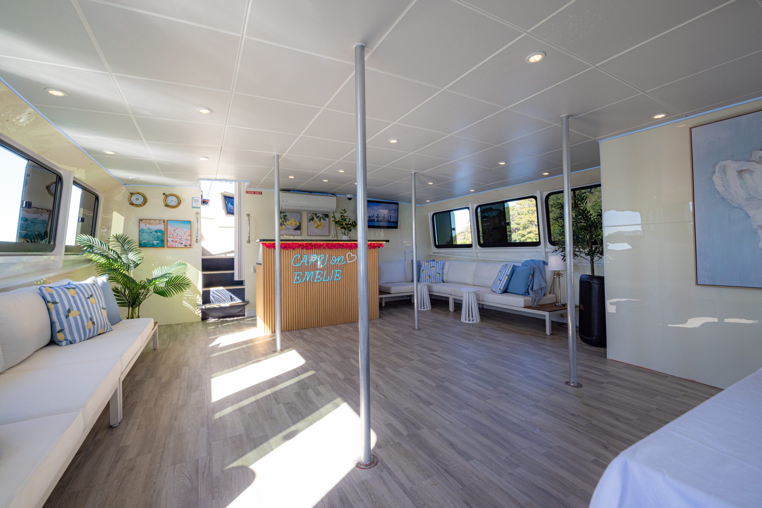 Spacious boat interior with white seating, wood floors, large windows, tropical plants, and a wooden counter labeled “Captain’s Club.” Bright natural light fills the modern, clean space.