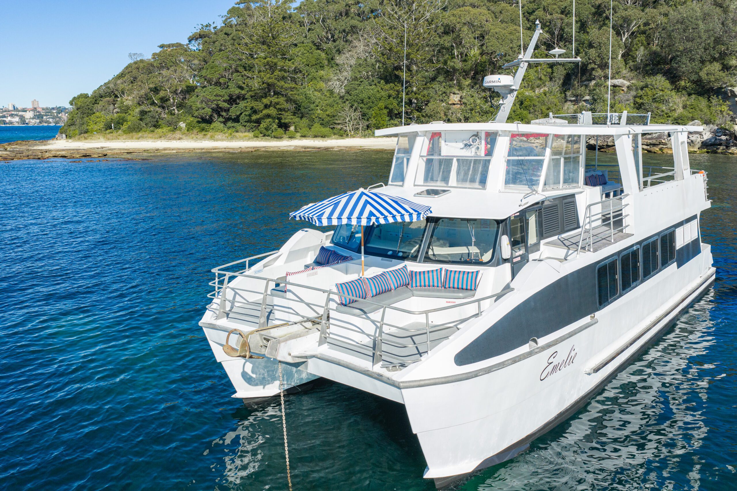 A white yacht named “Emilie” anchored near a green island, with striped blue lounge chairs and an umbrella on the deck, on clear blue water under a sunny sky.