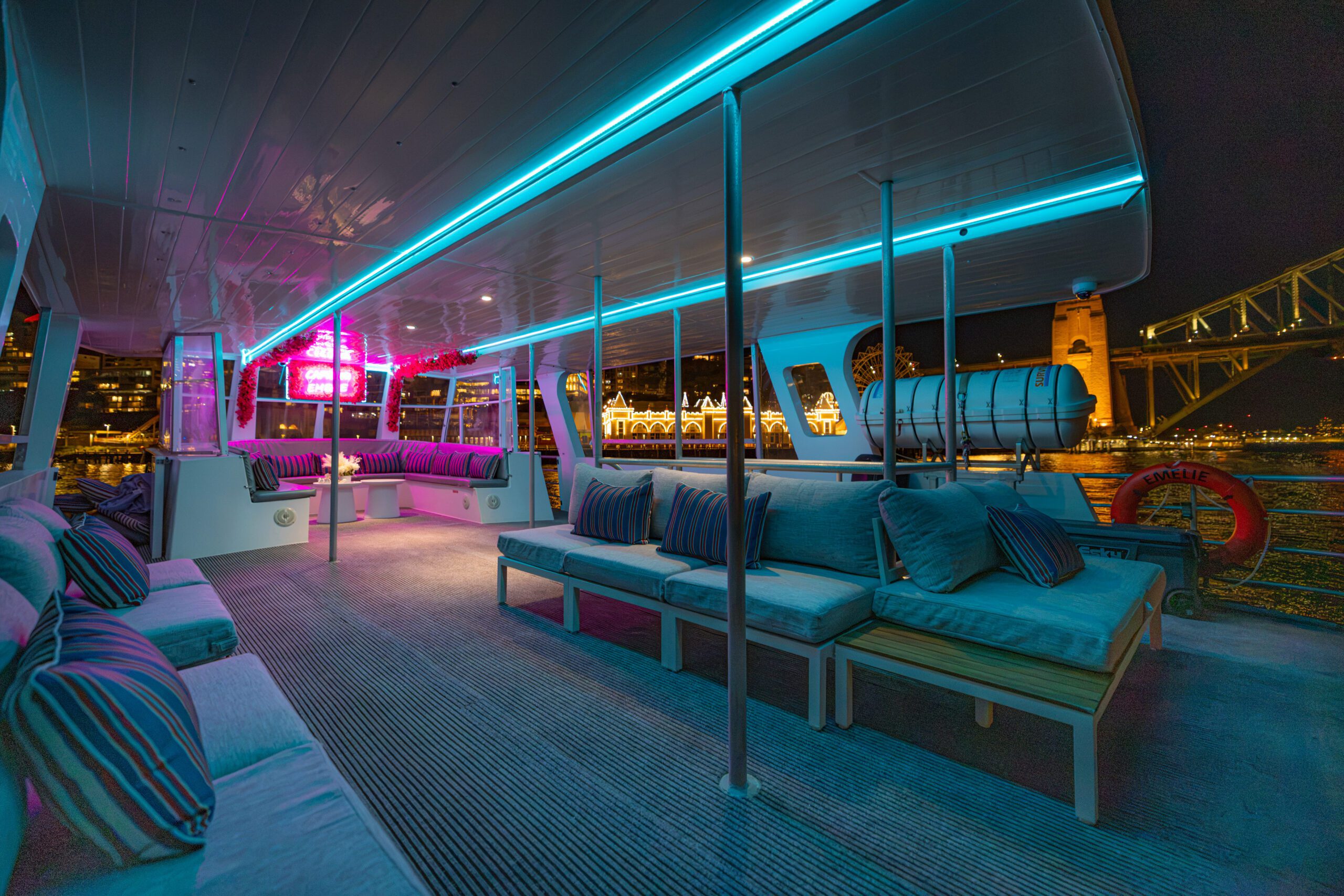 A modern boat deck at night with blue LED lights, cushioned seating, striped pillows, and views of a city skyline, bridge, and waterfront illuminated in the background.