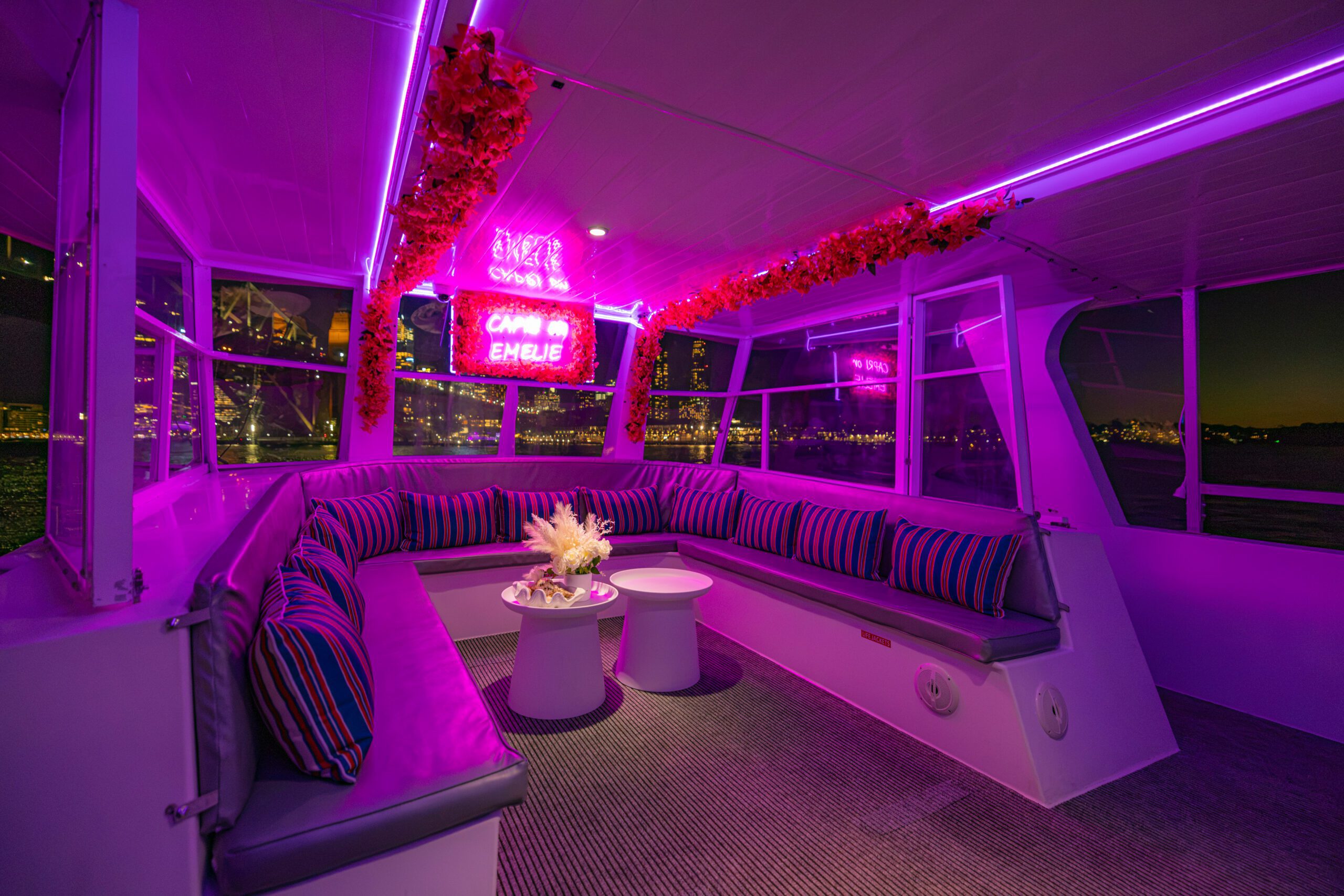 Interior of a boat with purple neon lights, striped cushions on curved benches, floral decorations, two small tables, and “Camp Emelie” neon sign. City lights are visible through the windows at night.