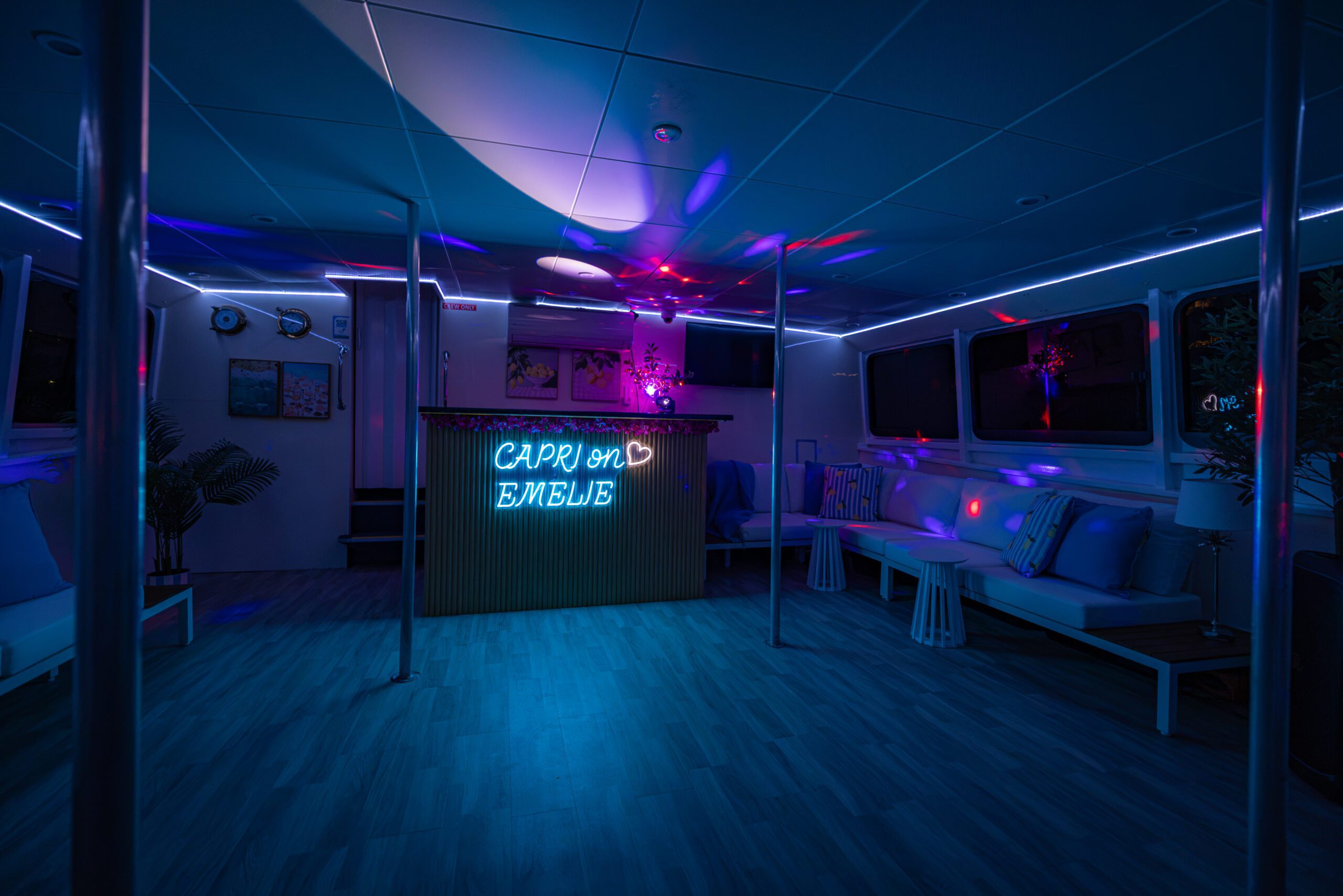 A lounge with blue and purple neon lights, a bar displaying a “CAPRI ON EMBLUE” sign, white seating, plants, stools, and large windows create a modern and colorful atmosphere.