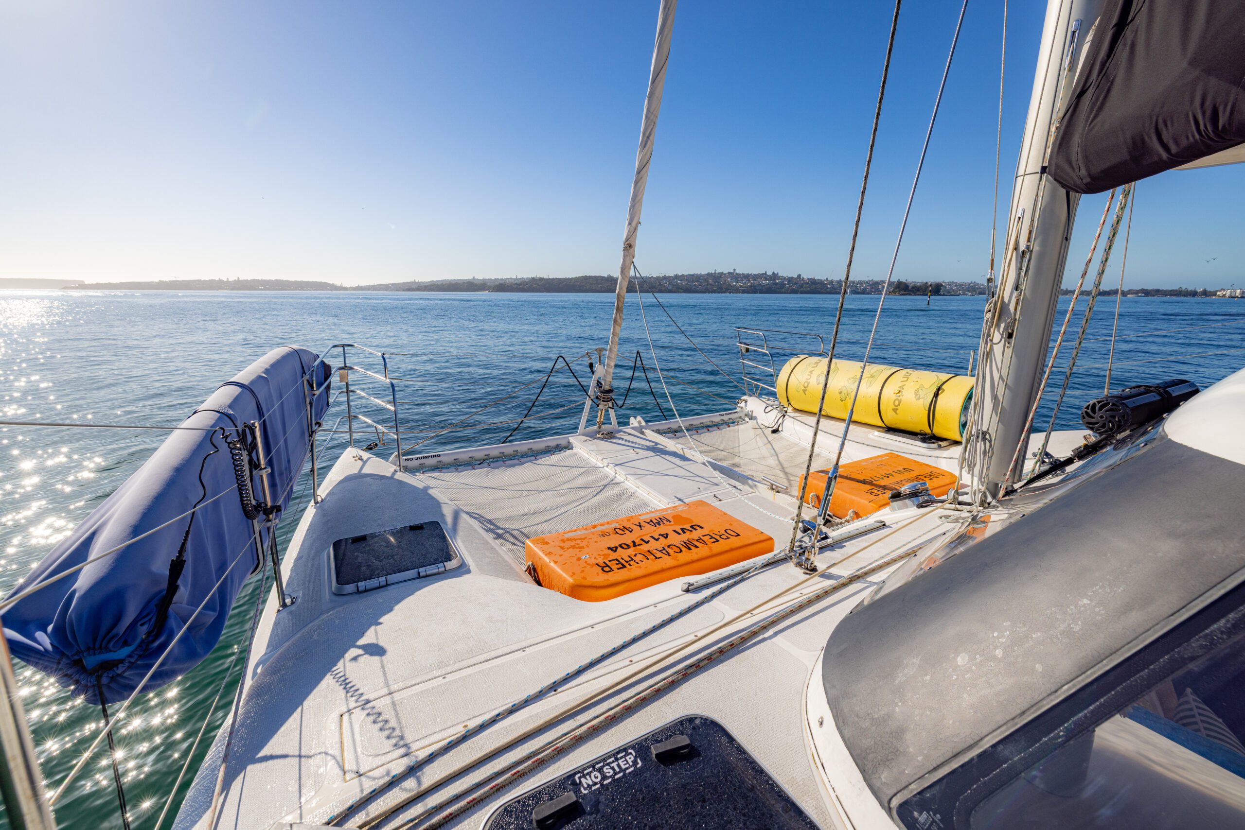 View from the deck of a sailboat on calm water, with bright sunlight, life rafts, a covered dinghy, and land visible in the distance under a clear blue sky.