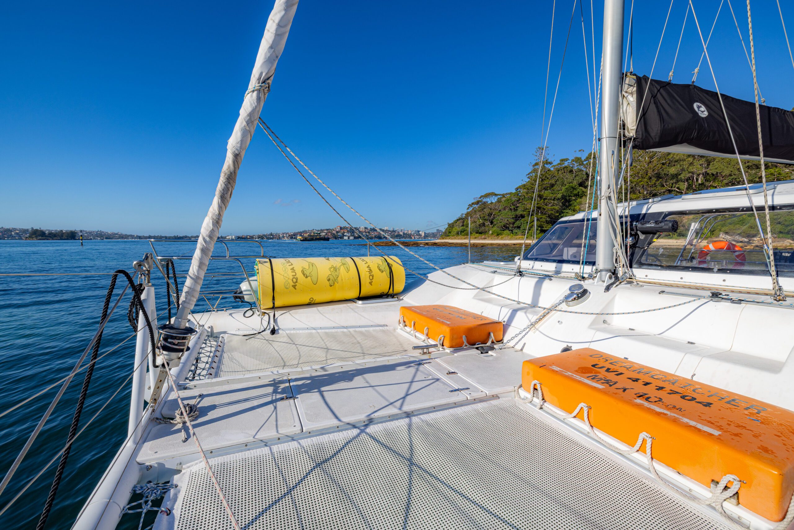 View from the deck of a white catamaran sailboat with orange and yellow storage containers, anchored near a green, forested coast under a clear blue sky.