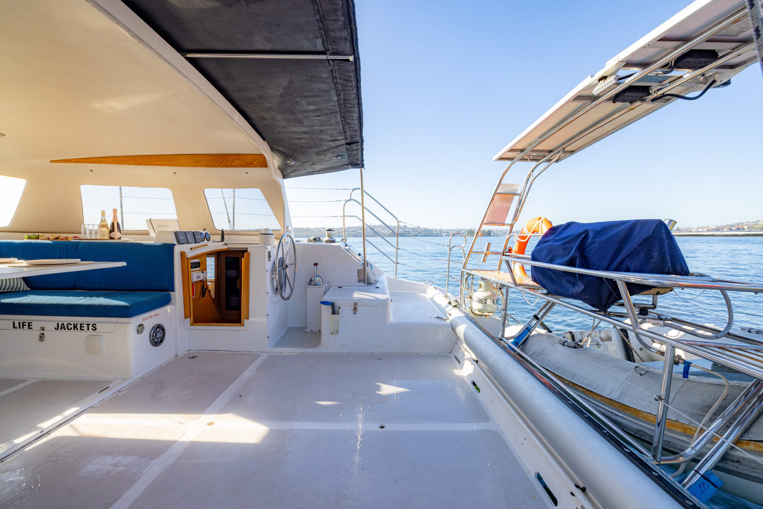 Open deck of a sailboat with seating area, life jackets storage, and a covered dinghy on the right, overlooking calm water and distant shore under a clear sky.