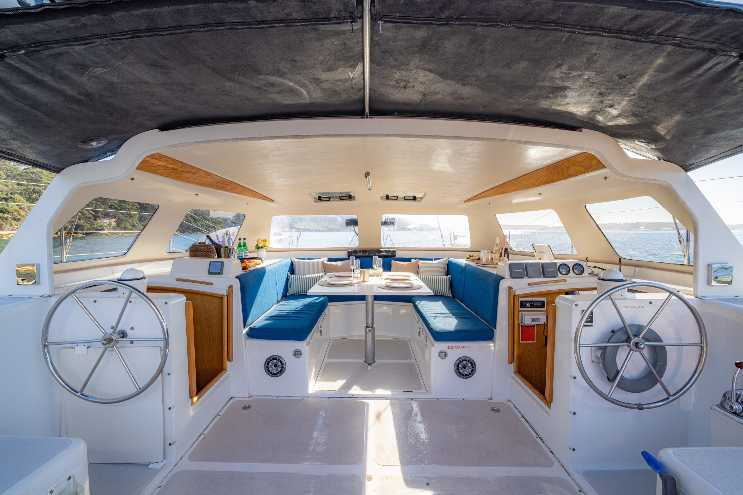The interior of a catamaran with two steering wheels, blue cushioned seating, dining tables, and large windows showing water and shoreline outside.