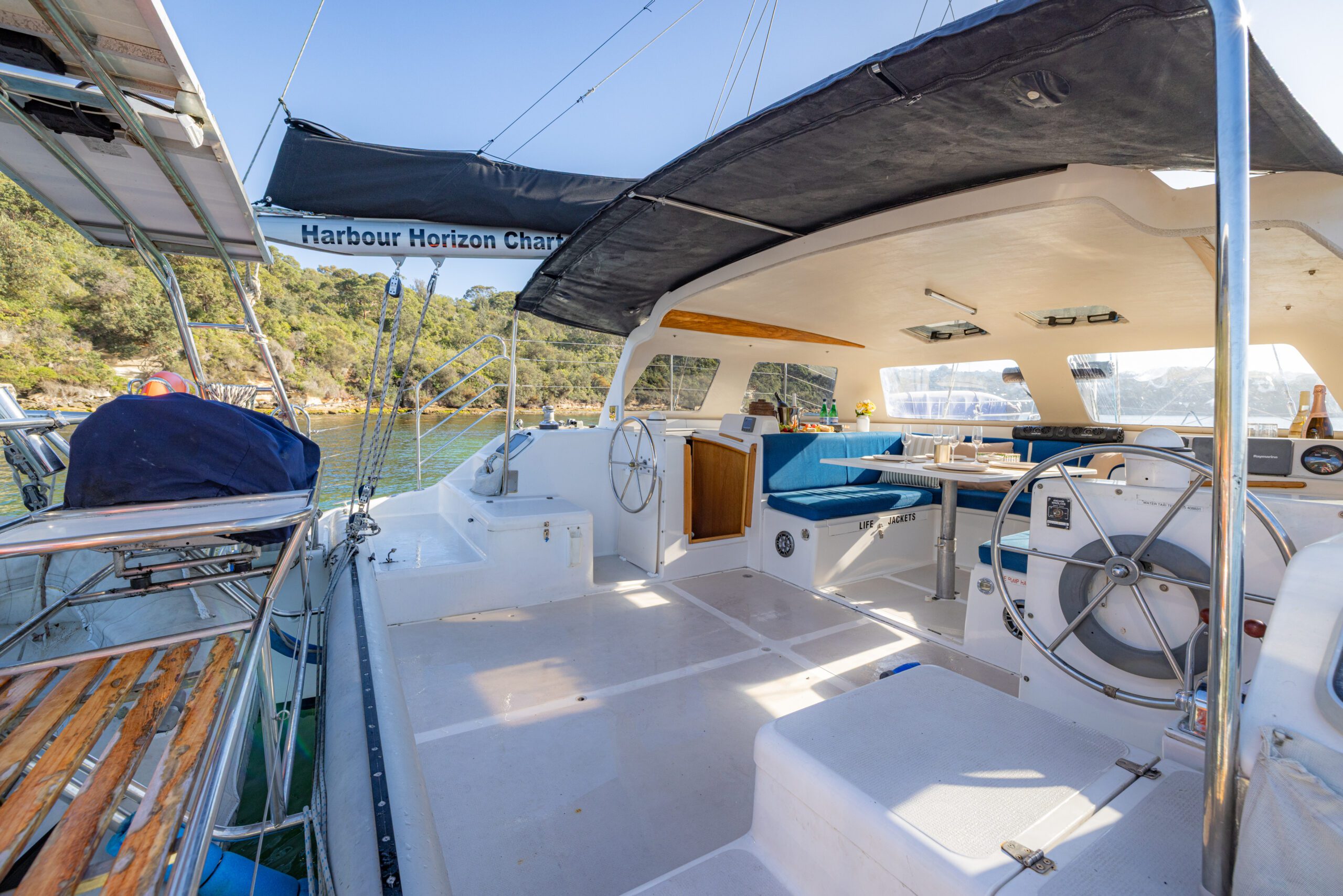 A spacious yacht deck with dual steering wheels, shaded seating area, and a dining table, docked near lush green shoreline under a clear sky.
