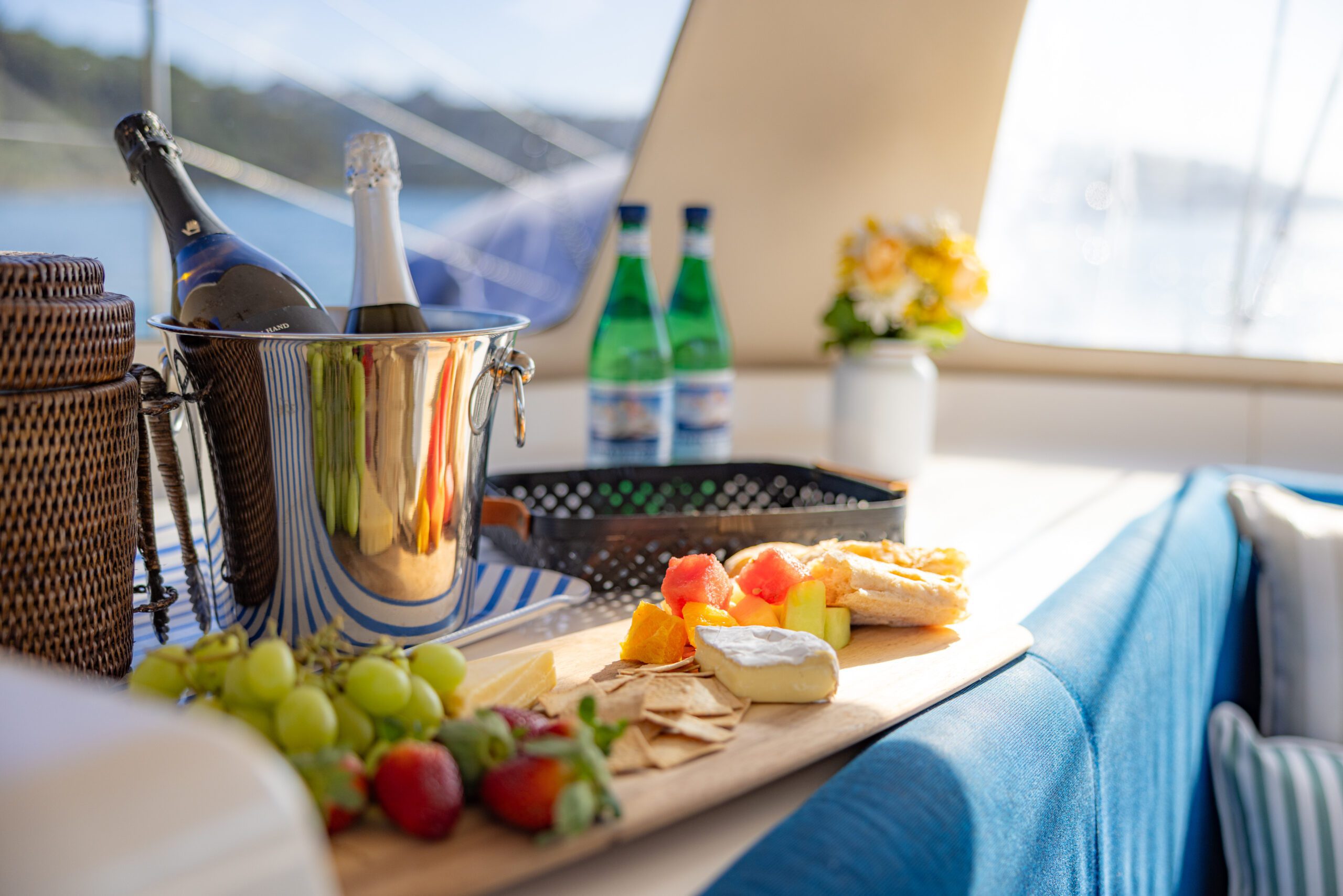 A wooden tray with fruit, cheese, and crackers sits on a yacht table beside bottles of water, champagne in a bucket, a picnic basket, and a vase with flowers. Sunlight streams through windows overlooking the water.