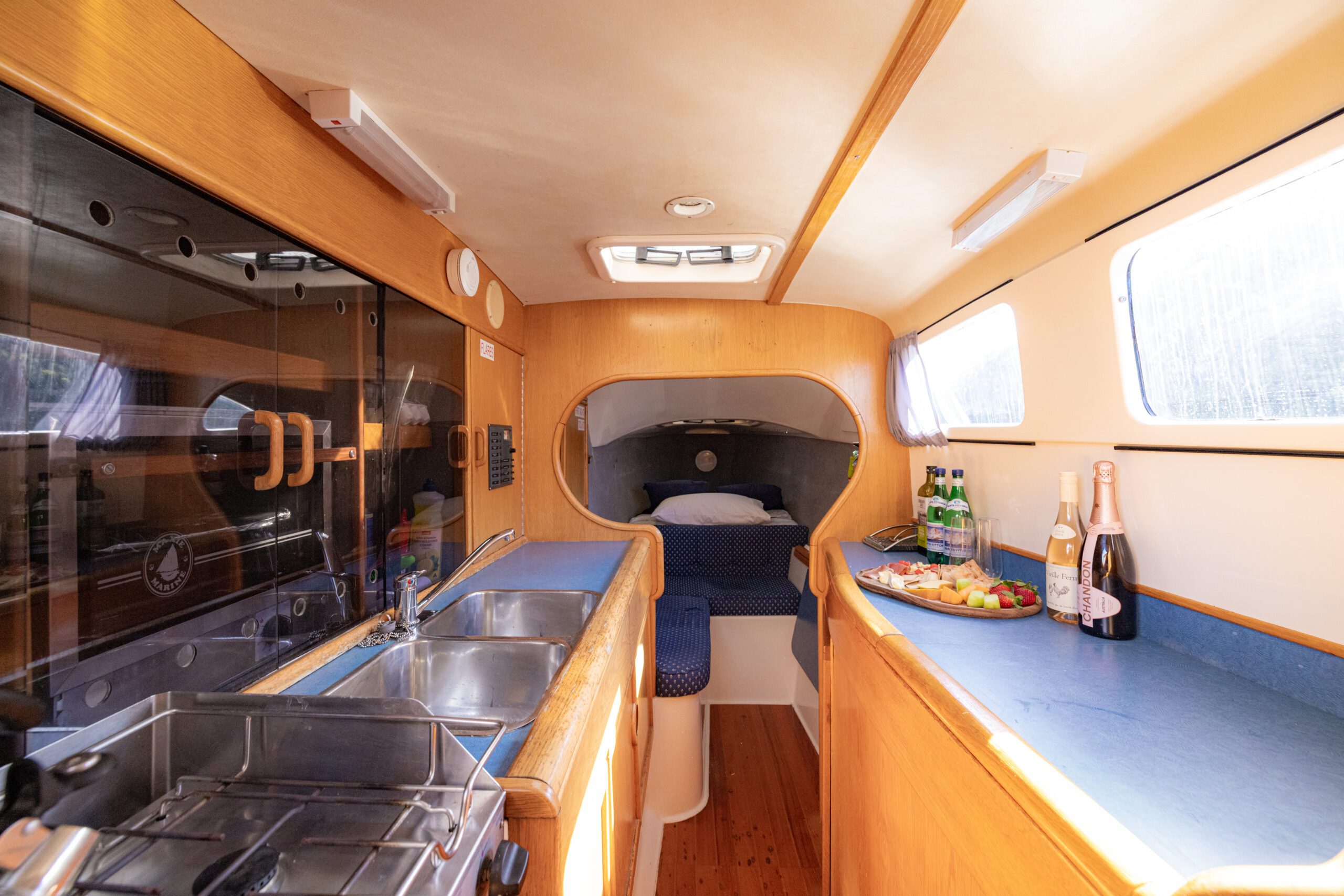 The interior of a boat cabin with a sink, stove, and counter on one side, and a bed in the sleeping area at the far end. A fruit platter and drinks sit on the counter by a sunlit window.