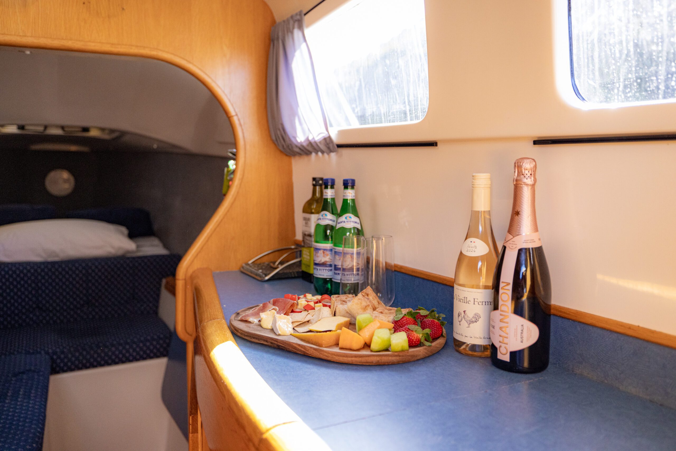 A wooden tray with assorted cheeses, fruits, and meats sits on a blue counter next to bottles of water, white wine, and champagne inside a cozy, sunlit boat cabin.