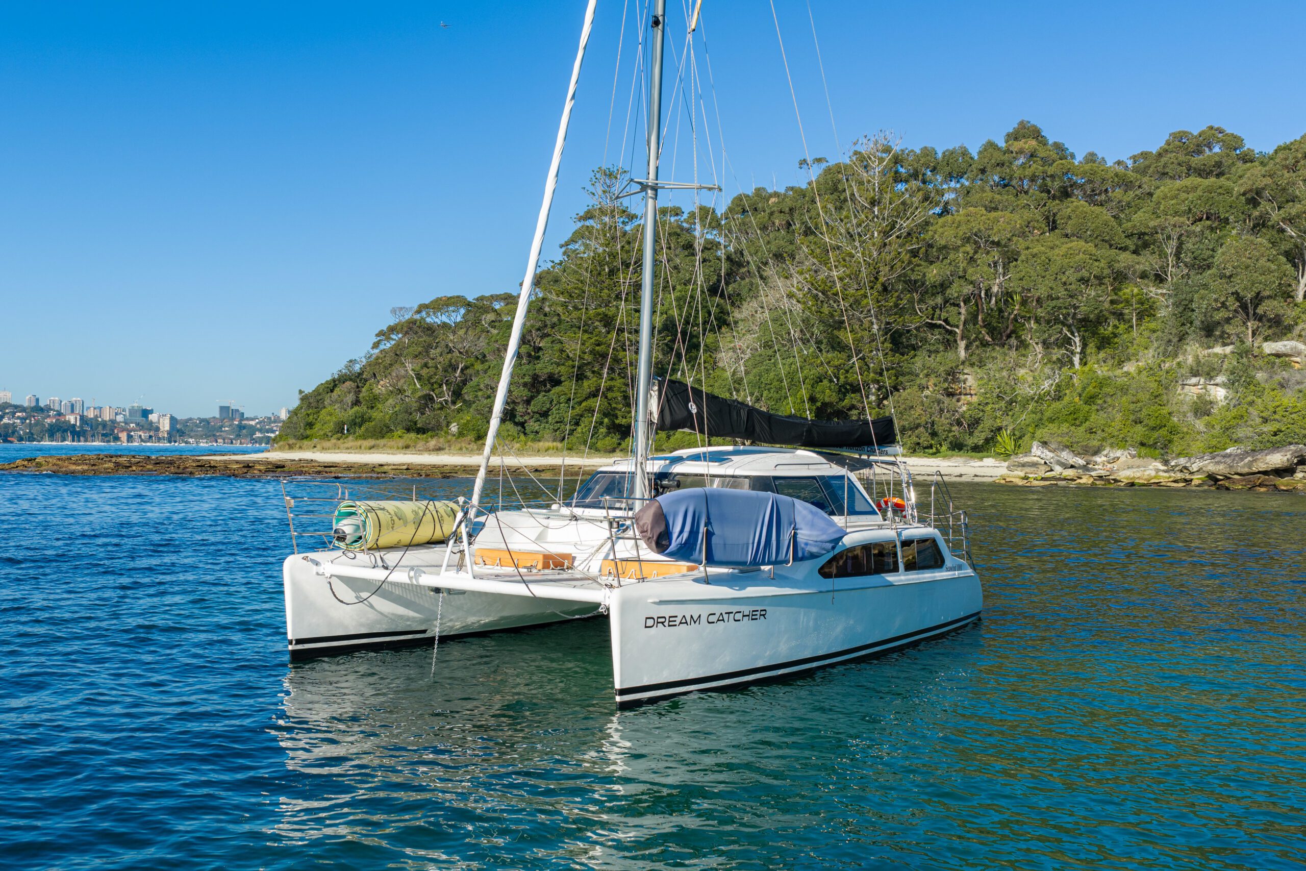 A white catamaran named “Dream Catcher” floats on calm blue water near a green, tree-lined shore under a clear blue sky.