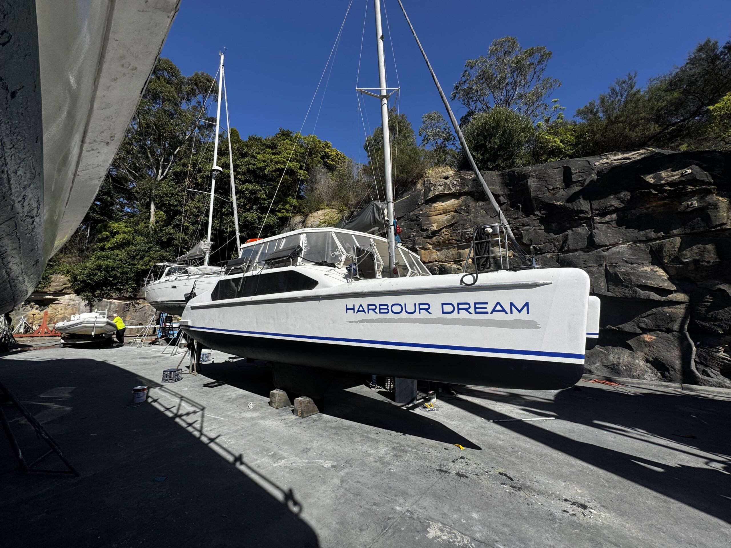 A white catamaran named “Harbour Dream” is on land, supported by stands, with rocky cliffs and trees in the background under a clear blue sky.