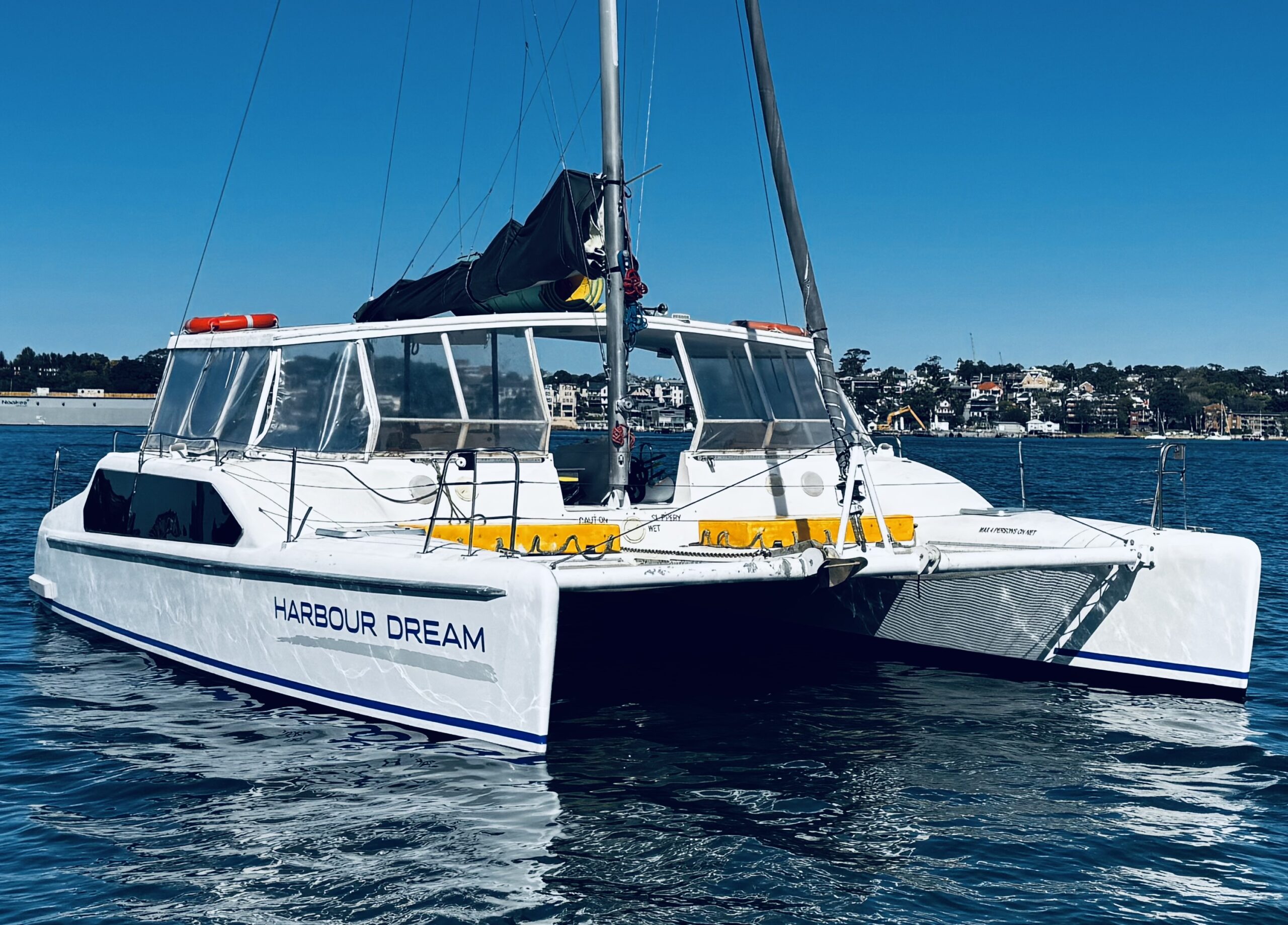 A white catamaran named “Harbour Dream” floats on calm water under a blue sky, with coastal buildings and trees visible in the distant background.