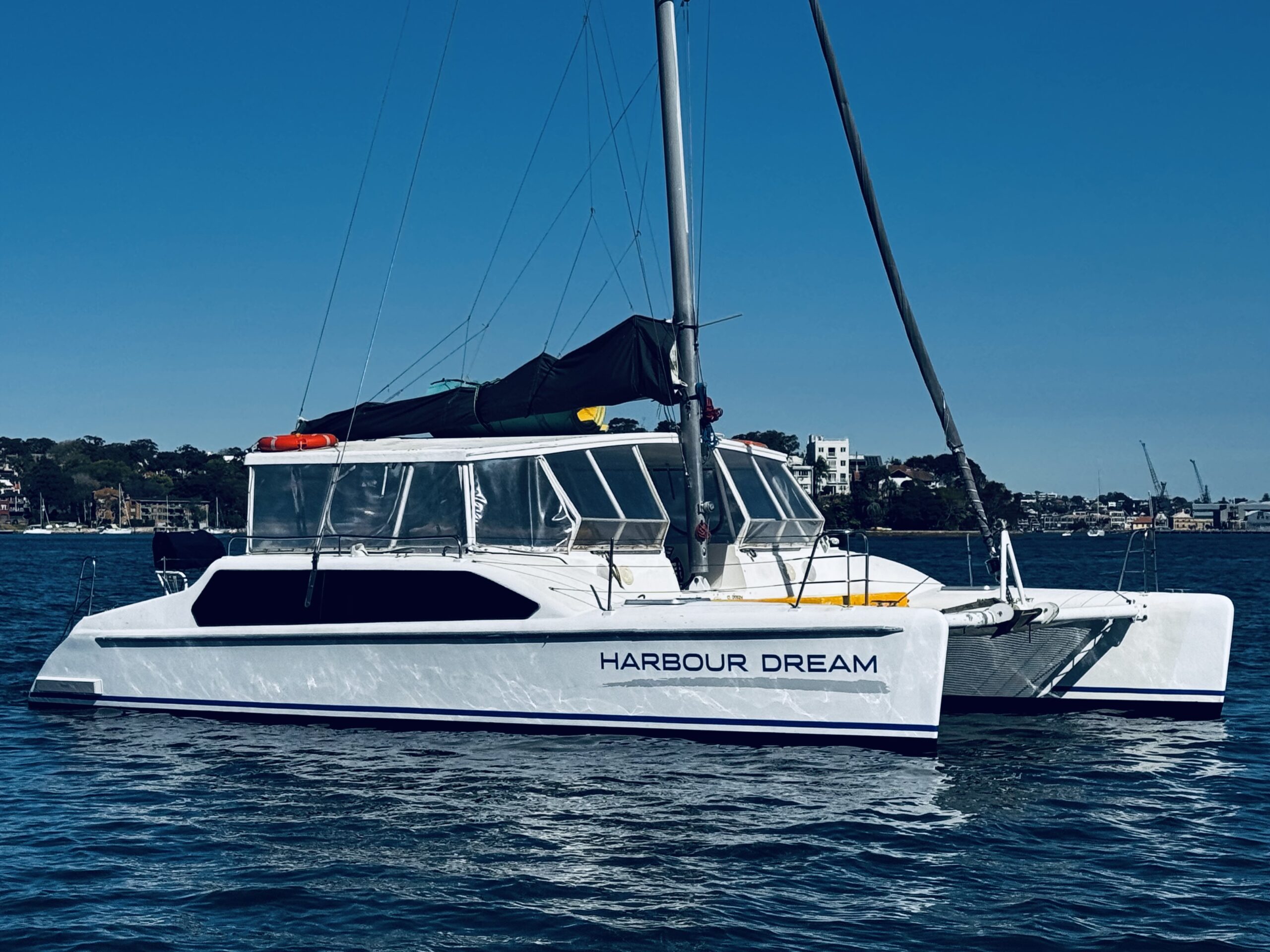 A white catamaran named “Harbour Dream” floats on calm water under a clear blue sky, with buildings and trees visible in the distant background.