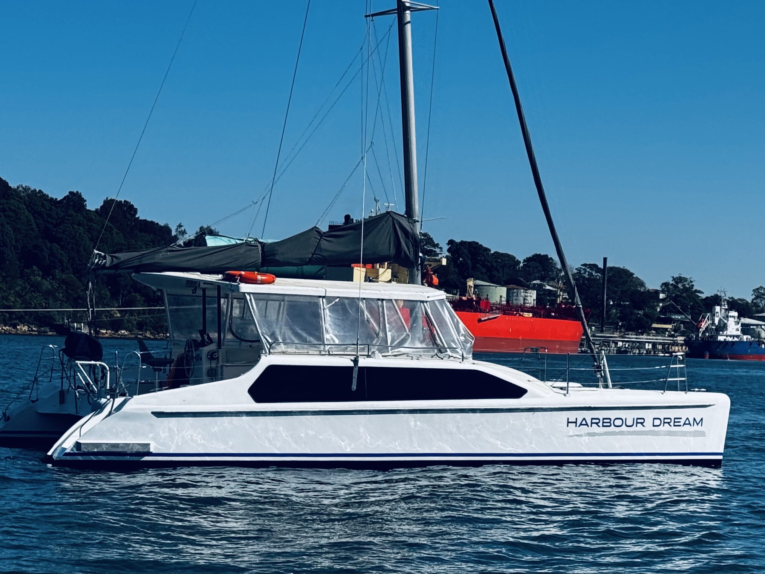 A white catamaran named “HARBOUR DREAM” floats on calm water with trees and a red ship in the background under a clear blue sky.