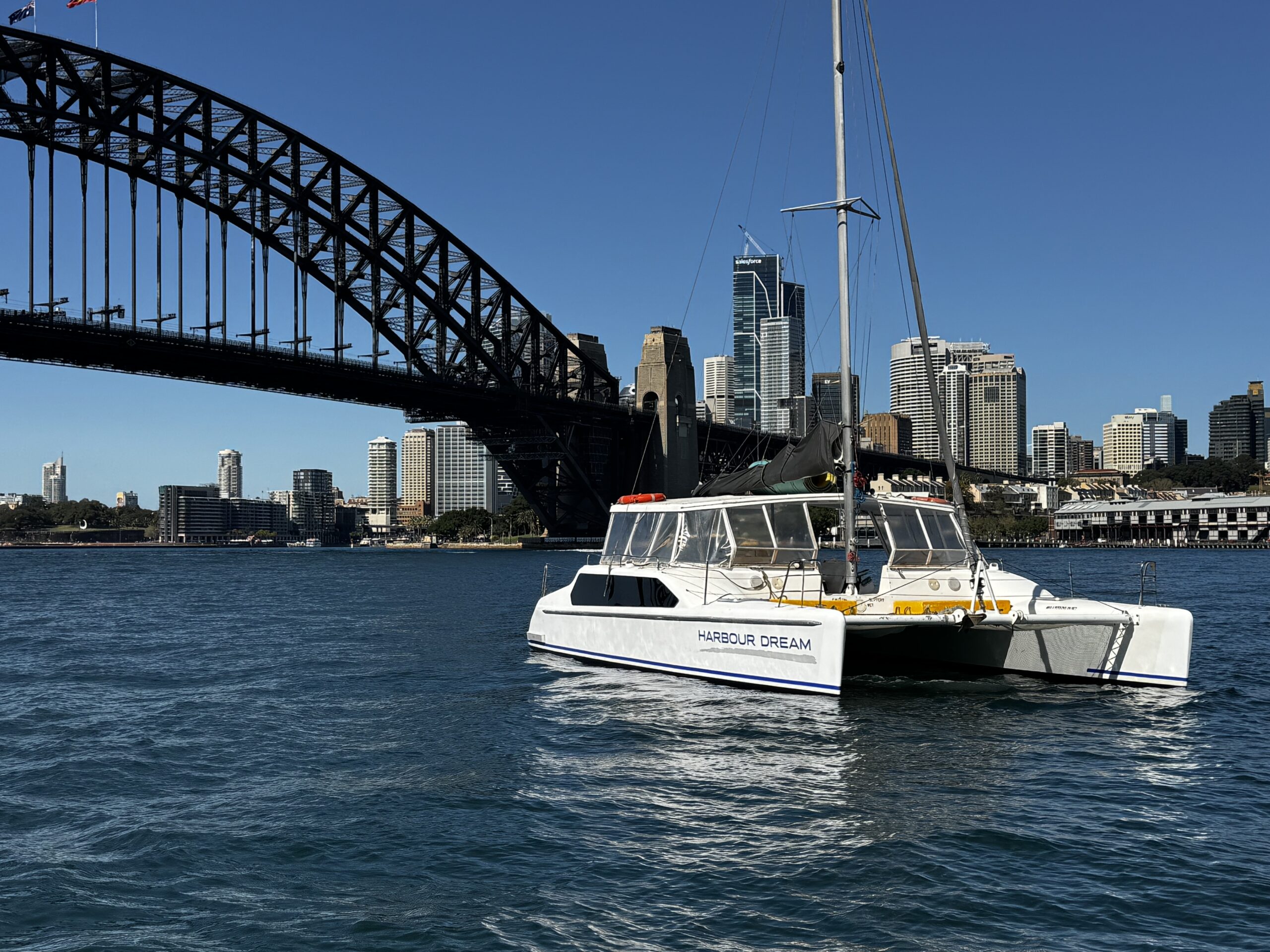 A white catamaran named “Harbour Dream” floats on blue water with Sydney Harbour Bridge and city skyline in the background under a clear blue sky.