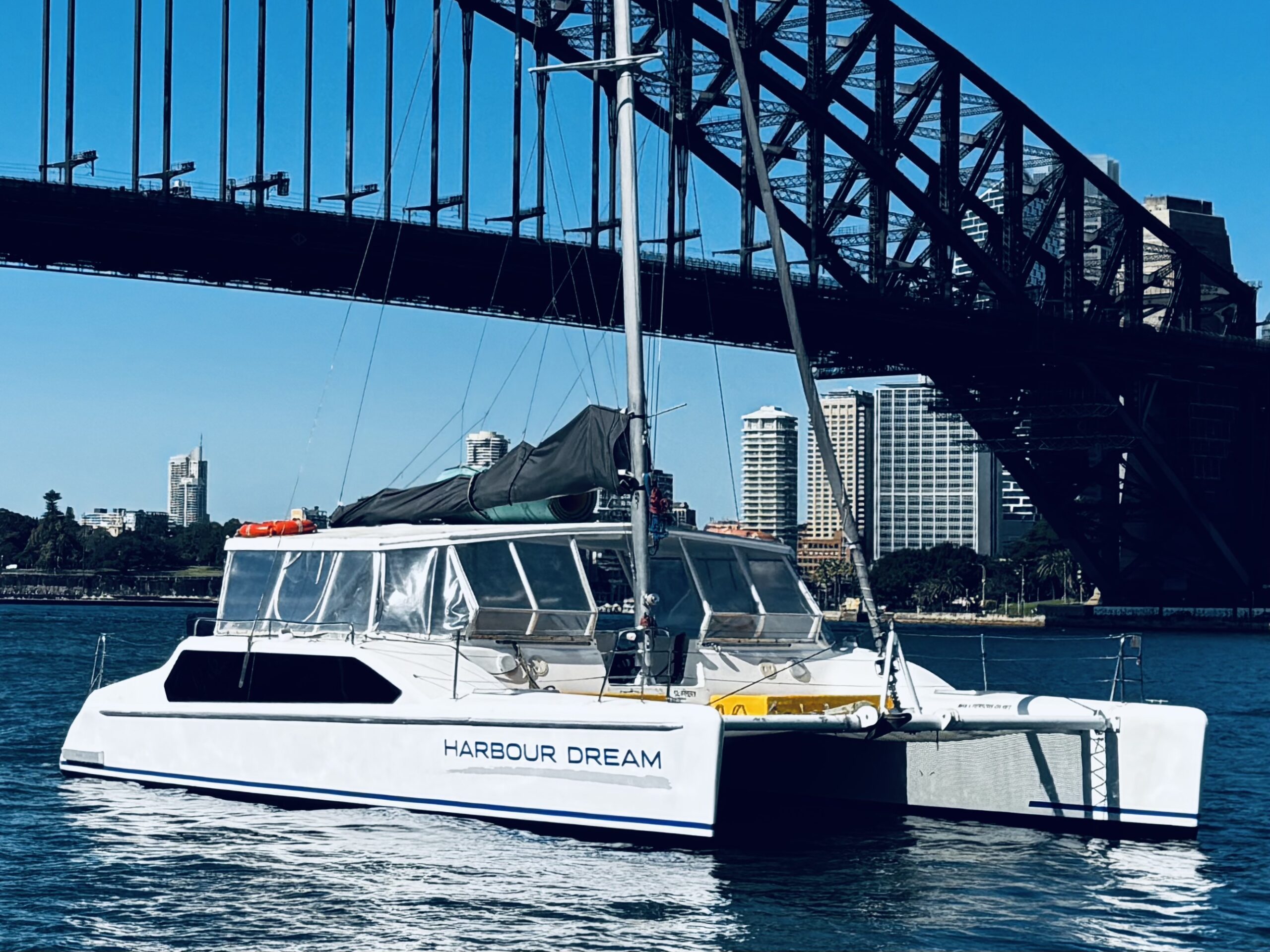 A white catamaran named “Harbour Dream” floats on water near a large steel bridge, with city buildings and a clear blue sky in the background.
