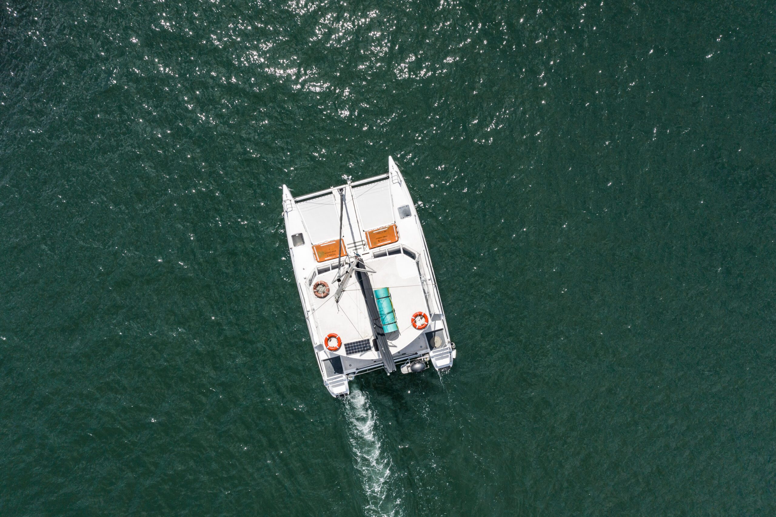 Aerial view of a white catamaran with orange seating and life rings, sailing on deep green water, leaving a visible wake behind.