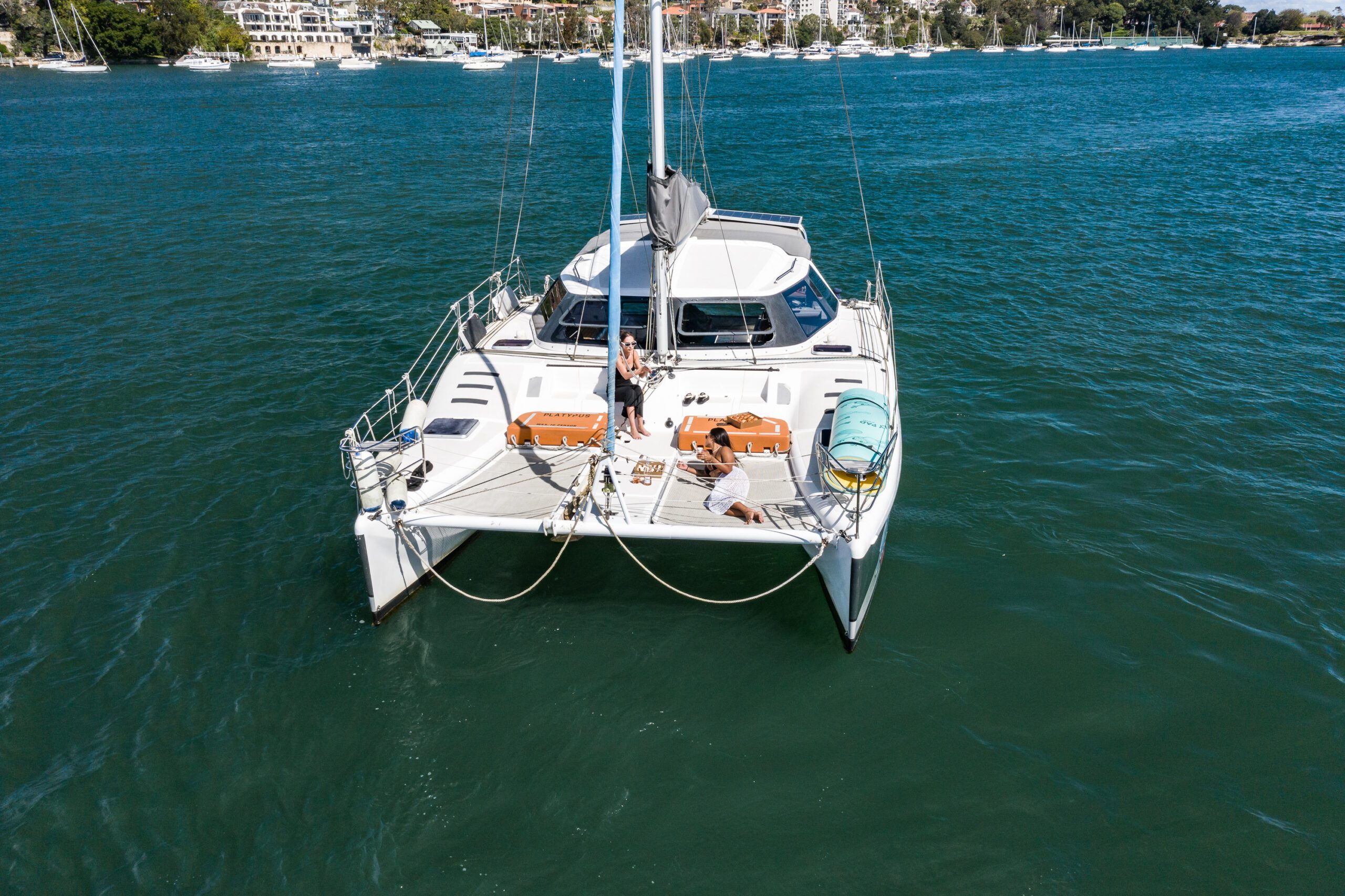 A catamaran with two people relaxing on the deck floats on calm blue-green water near a shoreline with boats and buildings in the background.