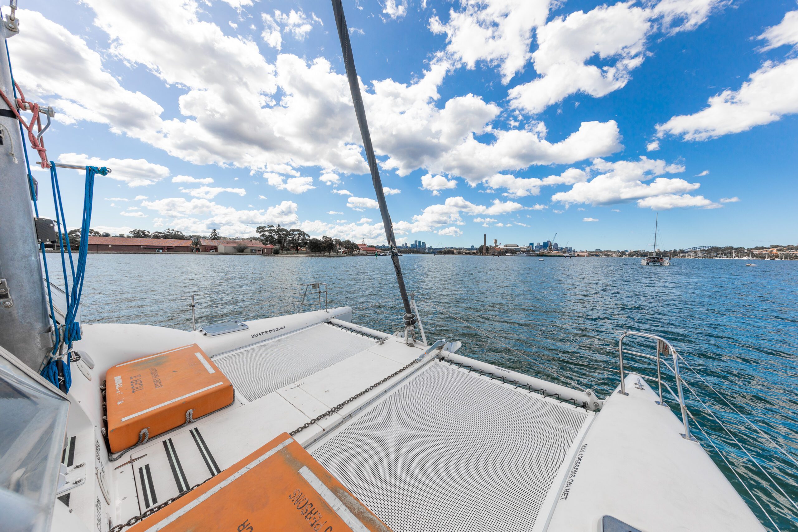 View from a catamaran deck on calm blue water, with orange storage containers, netted area, and cityscape and clouds in the background under a bright sky.