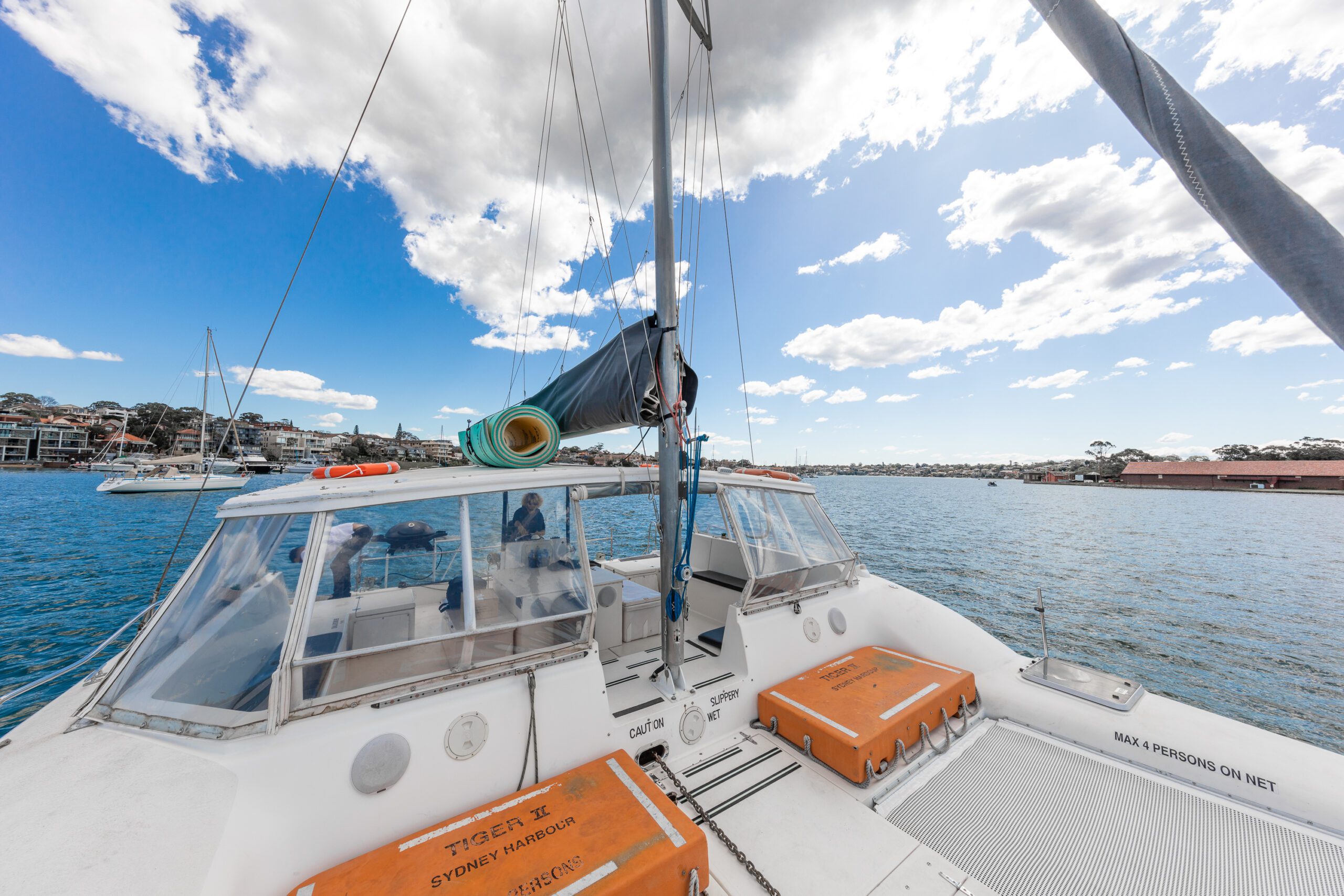 A catamaran with orange life rafts and equipment on deck is docked on calm blue water under a bright sky with scattered clouds, with a shoreline and buildings in the background.